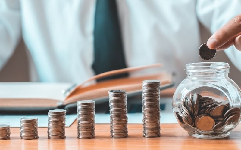 Photo of coins arranged like towers on a table with a man holding a coin and about to place in a jar filled with coins