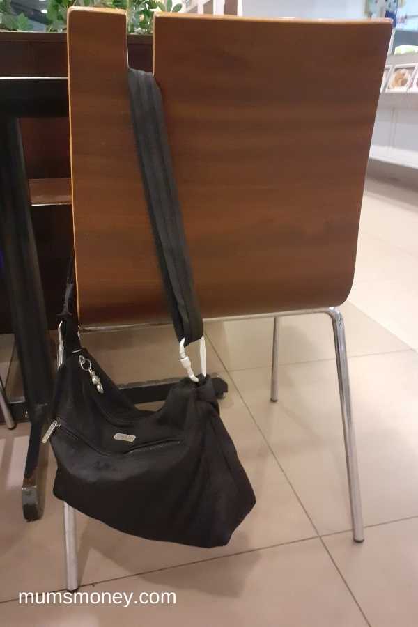 Black crossbody handbag hanging from the back of a brown wooden chair in a foodcourt.