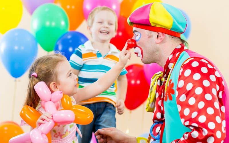 Photo of a boy, a girl and a clown where the girl is playfully touching the nose of the clown with many colorful balloons