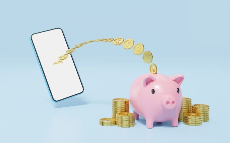Image of a cellular phone with coins around the pink piggybank in blue background.